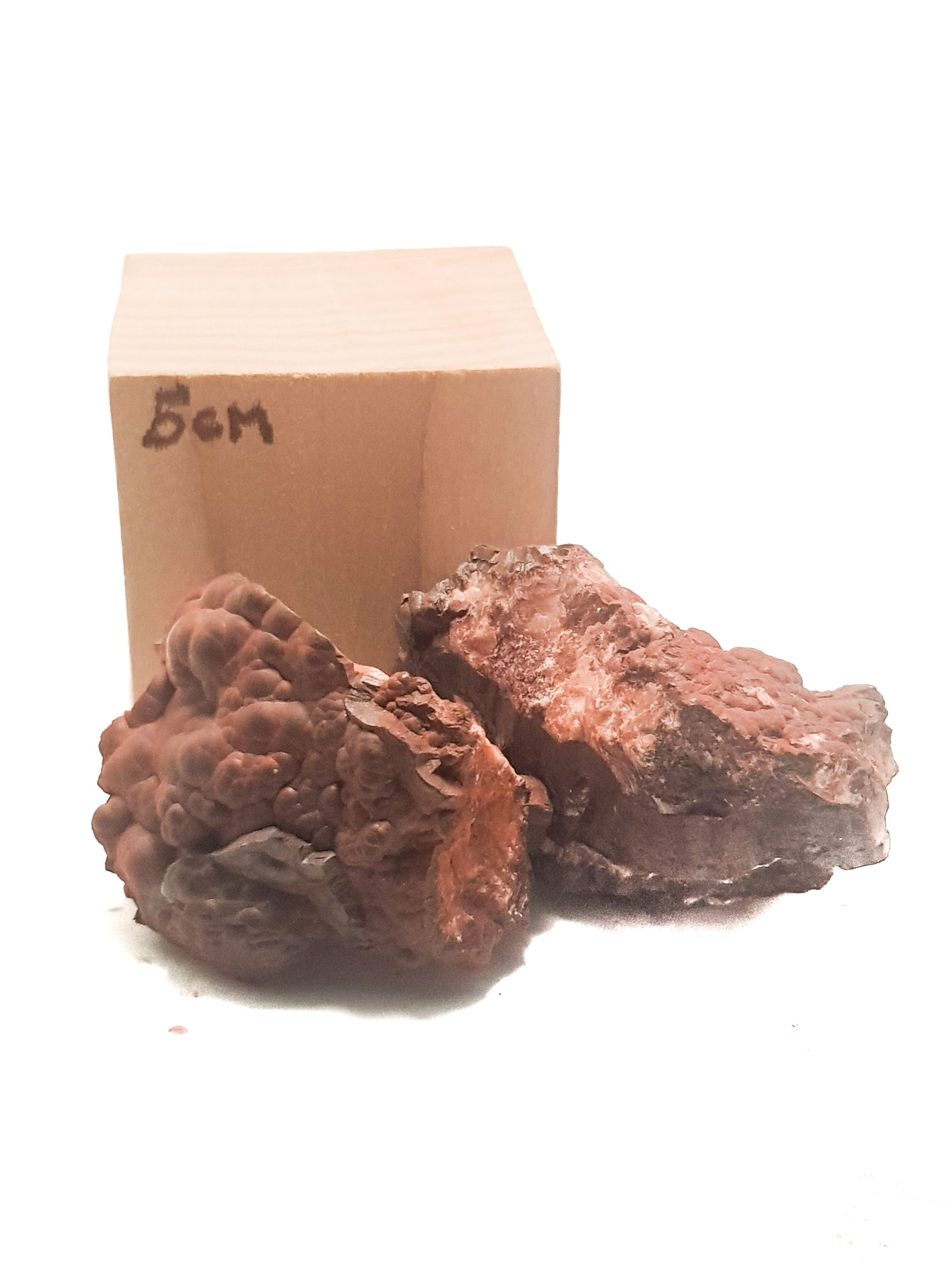 two samples of raw bottroydial hematite. One sample shows the bubbly surface, the other sample shows that the bubbles are layered when cut through. The samples are not to a wooden cube which states that it is 5cm. It is given for scale.