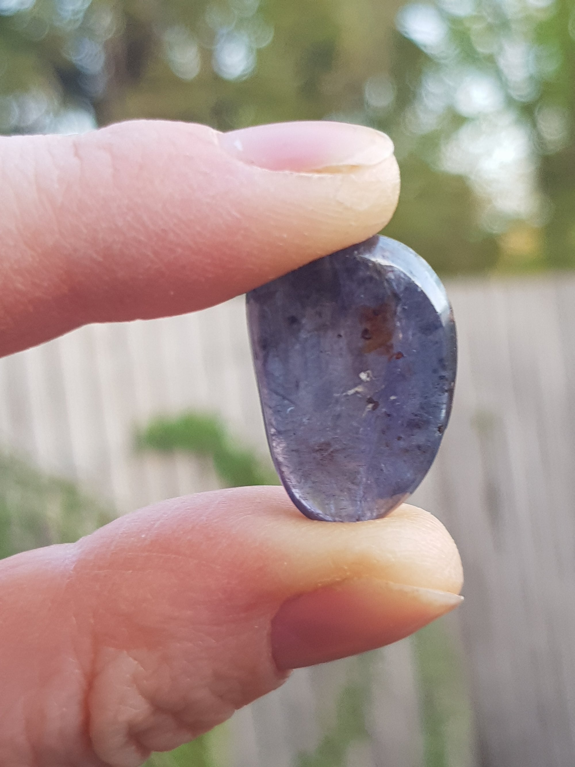 A small polished sample of translucent polite. It is held up to the light. It is a  violet grey. It has small inclusions of an orange mineral and of a black mineral.