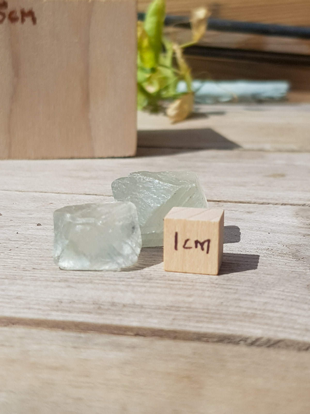 Two pale green fluorite octohedra on a wooden bench, they are next to a 1cm cube for scale. They are approximately 1.4cm cubed
