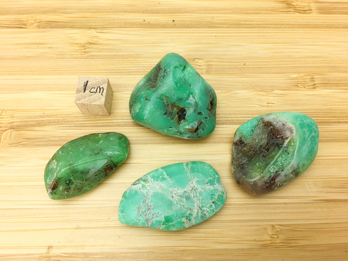 four pieces of apple green chrysoprase on a wood grain surface. A 1cm cube is placed next to one of the samples for scale. The samples are approximately 2cm long