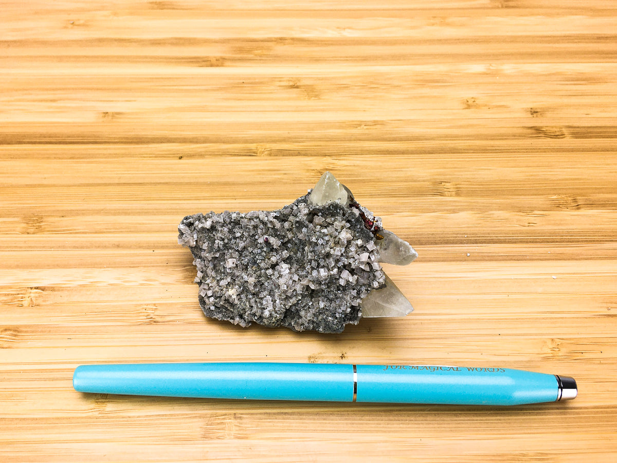 This shows a sample of rhombic calcite crystals which have been placed against a fountain pen for  size comparison. The calcite crystals are very small, but well formed. the points of 3 dogtooth calcite crystals are also visible in this sample.