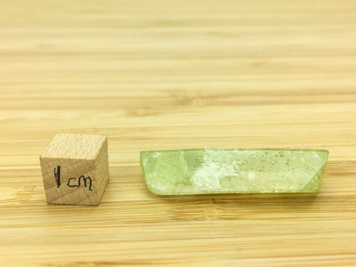 prismatic brazilianite crystal on a good grain surface. The crystal is prismatic and well formed. It is  green. It is gemmy. The crystal is placed next to a 1cm cube for scale. The crystal is 3.5cm long