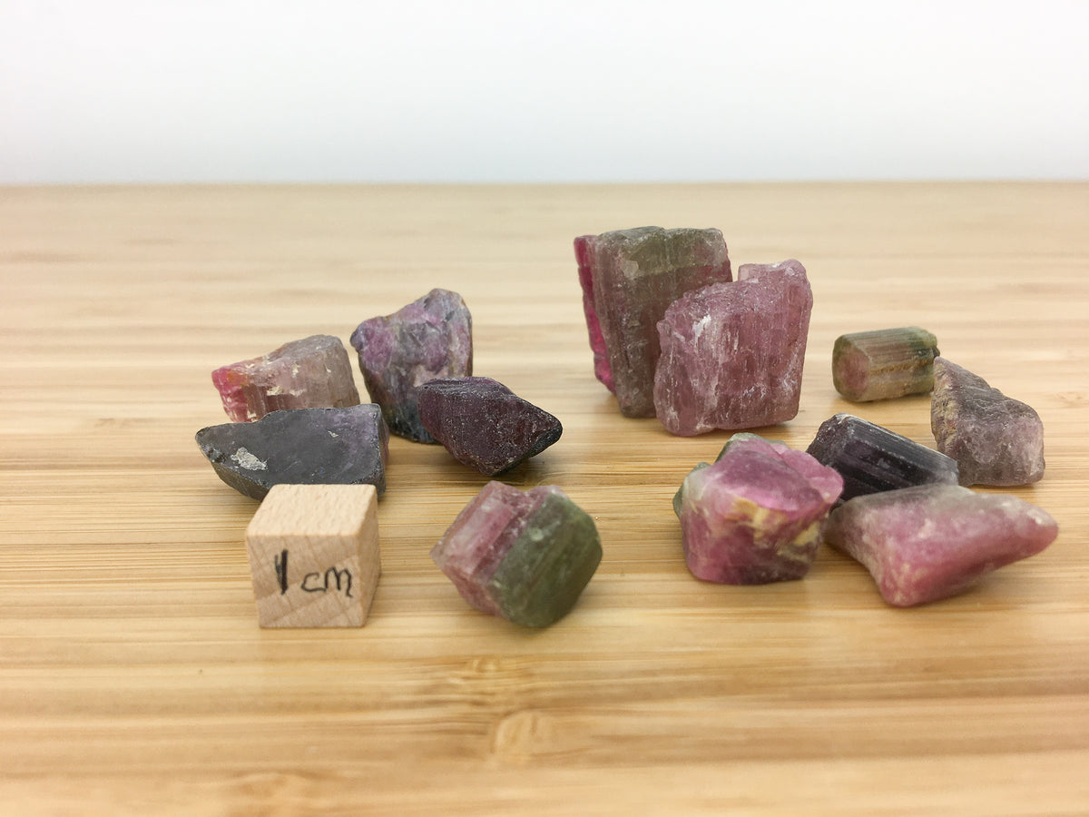 well formed elbaite (tourmaline) crystals next to a 1cm cube for scale. They are all about 1cm high and just under 1cm wide. They are pink and green and show concentric zoning.