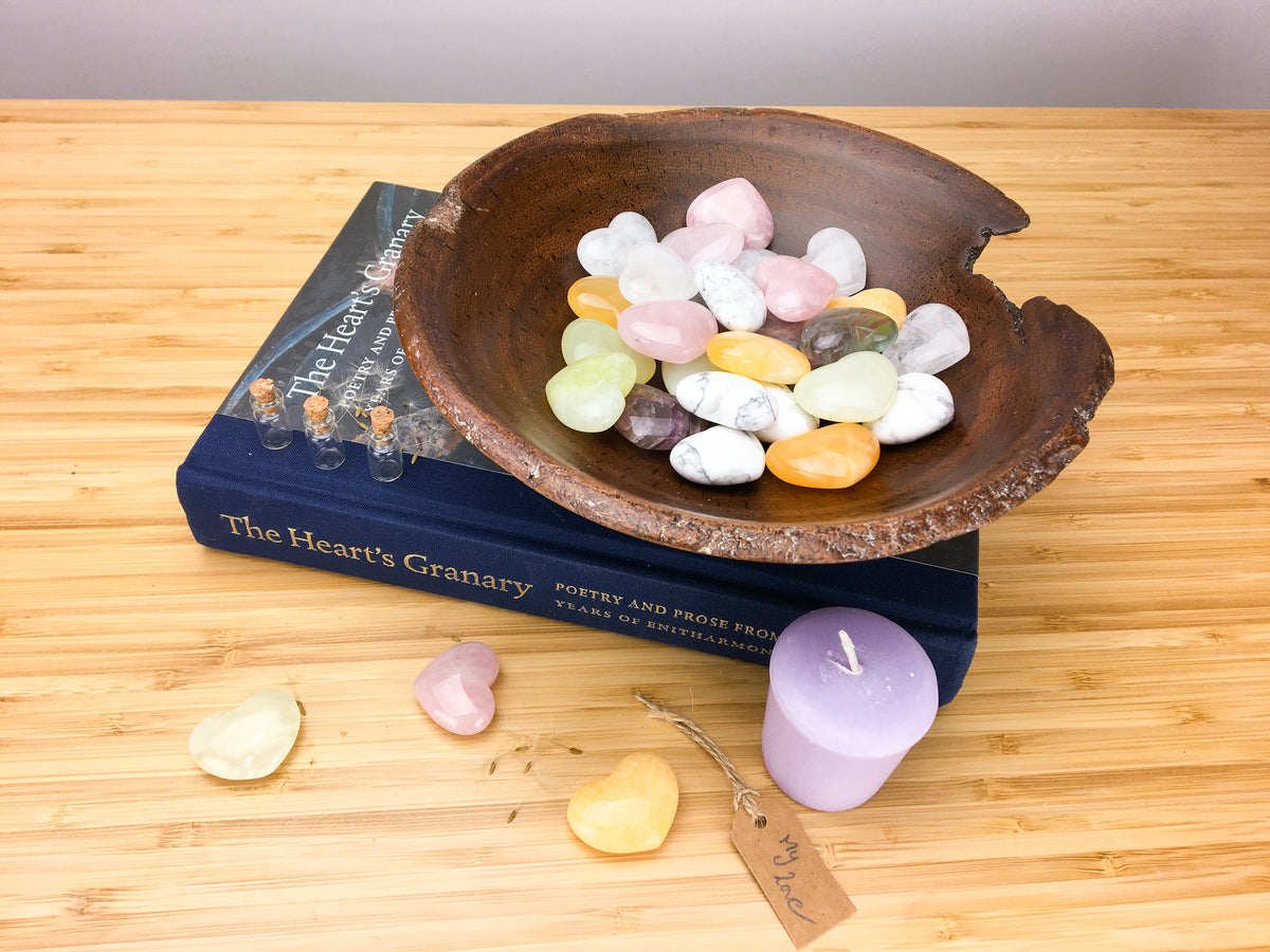 A hand carved wooden bowl filled with pastel coloured carved stone hearts. The bowl stands on top of a book called the hearts granary. The book is sitting on a light wood grained surface. A purple candle, three hearts and a table saying my love is also lying on this surface.