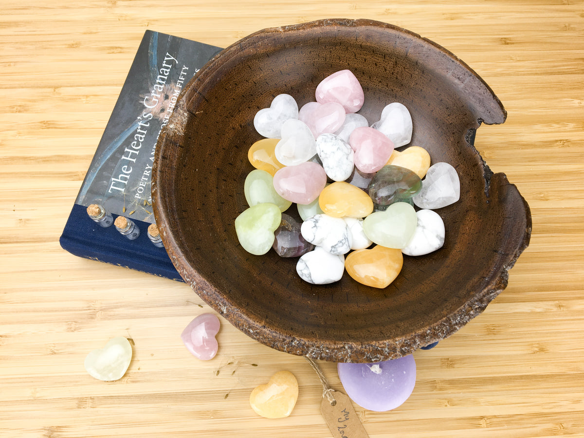 A hand carved wooden bowl filled with pastel coloured carved stone hearts. The bowl stands on top of a book called the hearts granary. The book is sitting on a light wood grained surface. A purple candle, three hearts and a lable saying &quot;my love&quot; is also lying on this surface.