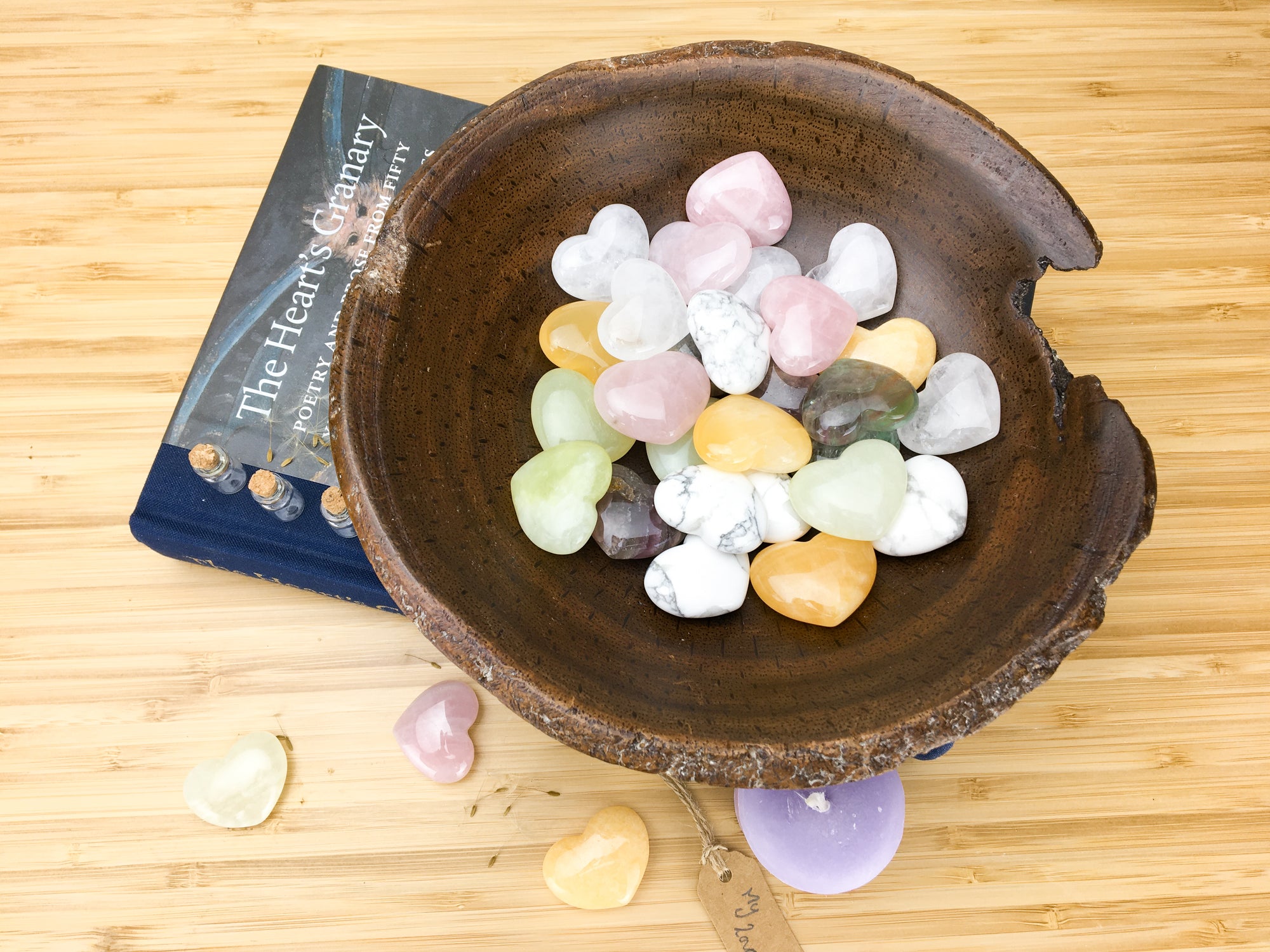 A hand carved wooden bowl filled with pastel coloured carved stone hearts. The bowl stands on top of a book called the hearts granary. The book is sitting on a light wood grained surface. A purple candle, three hearts and a lable saying "my love" is also lying on this surface.