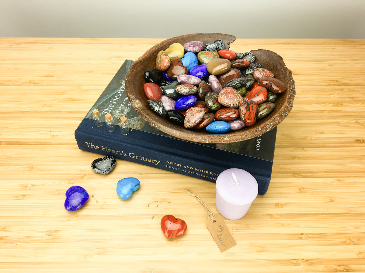A hand carved wooden bowl filled with brightly coloured carved stone hearts. The bowl stands on top of a book called the hearts granary. The book is sitting on a light wood grained surface. A purple candle, four hearts and a lable saying my love is also lying on this surface.
