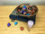  A hand catved wooden bowl containing a mixture of darkly coloured crystal hearts. The bowl is sitting on too of a book called the hearts granary. The book is on a wood grained surface. There is a purple candle and three coloured hearts on this surface