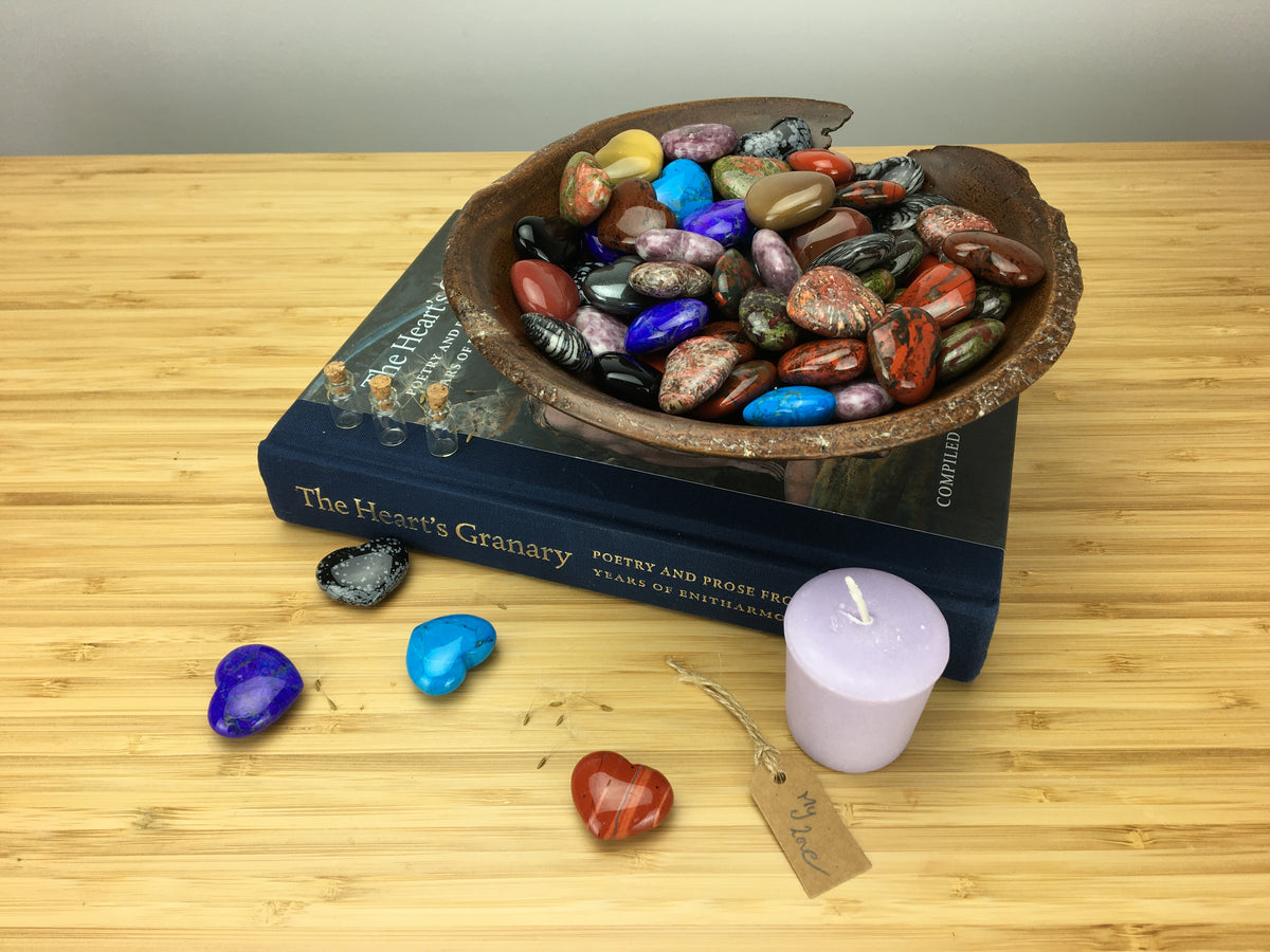 A hand carved wooden bowl is full of multicoloured crystsl hearts. The bowl sits on top of z book cslled the hearts granary. The book sits on a grained surface. A purple candle and foyr coloured hearts are on the grai ed surfacd. There is slso s hand written label which says I love you