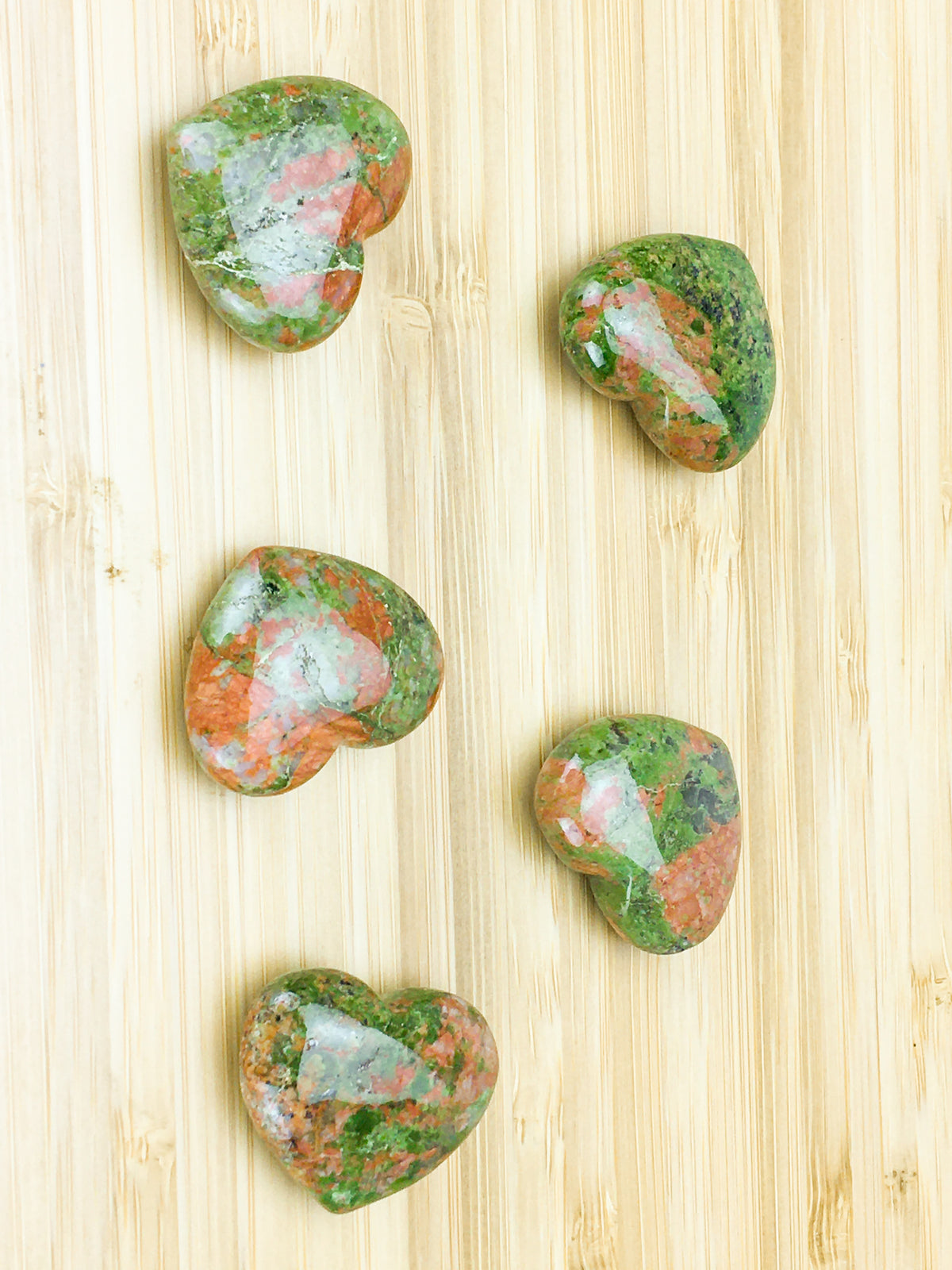 Five unakite hearts on a pale wood grain surface.