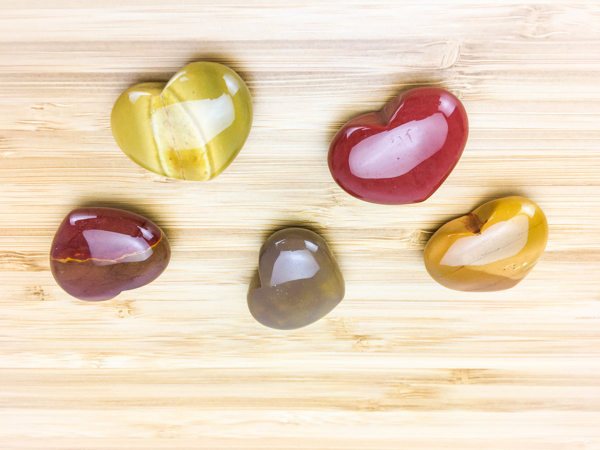 Five mookaite hearts on a grained wood table. The moojaite shows intense colour variation. Some pieces are red and some are orange 
