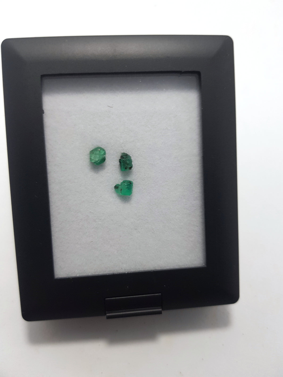 3 raw emerlds in a display case. The display box is black, the emeralds are displayed on white foam.