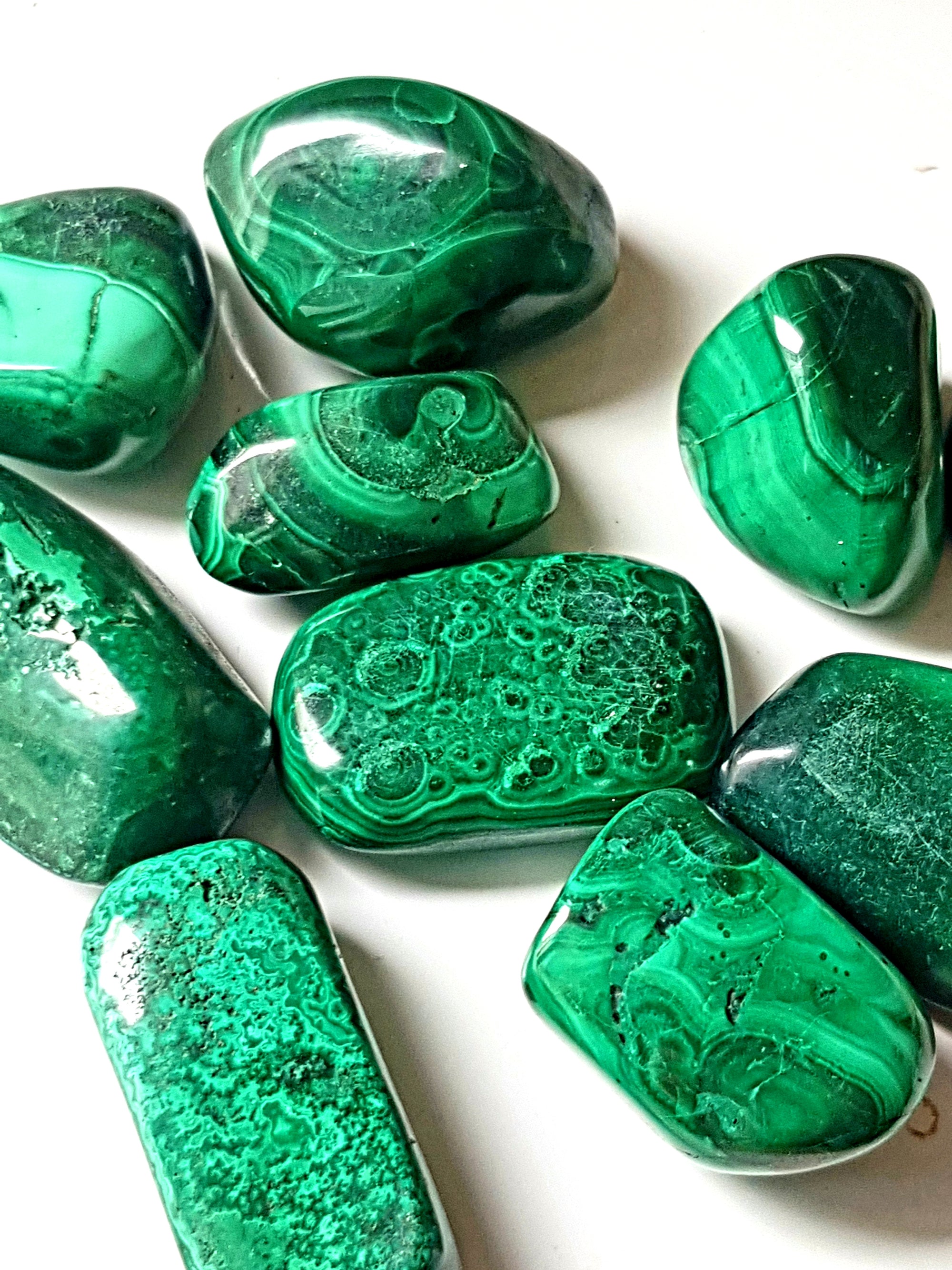 nine tumbled pieces of malachite. the pieces are smooth and elongate. They are banded and vary in colour from green to dark green. Some of the bands are patterned and look like little eyes.