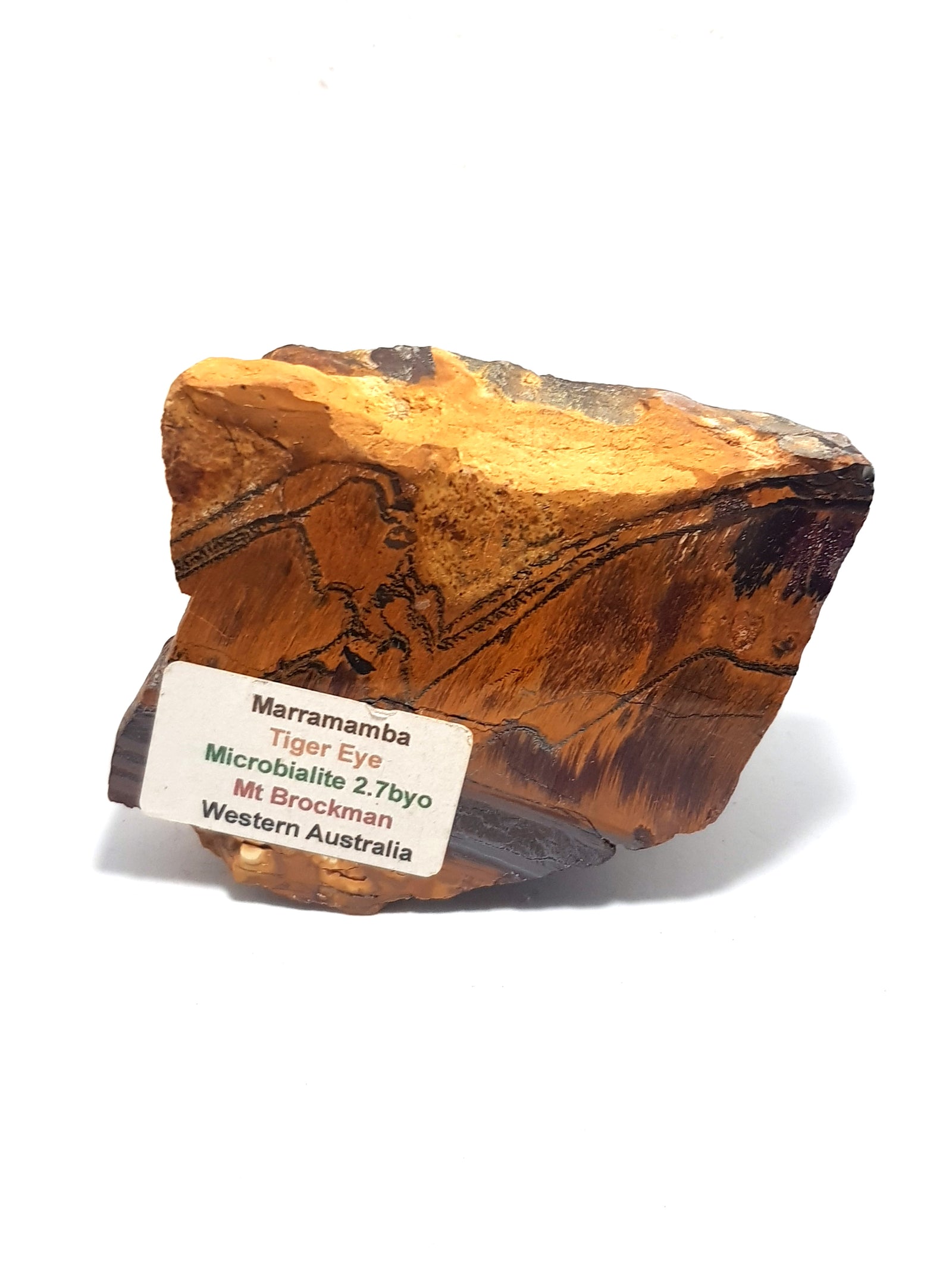 marramamaba tiger eye. this is an irregularly shaped chunk of raw rock. it is orange and displays chatoyancy. it contains thin zigzag bands of grey iron. there is a thicker band at the bottom of the rock. the rock has a printed label giving geological details. these details are replicated in the main entry.