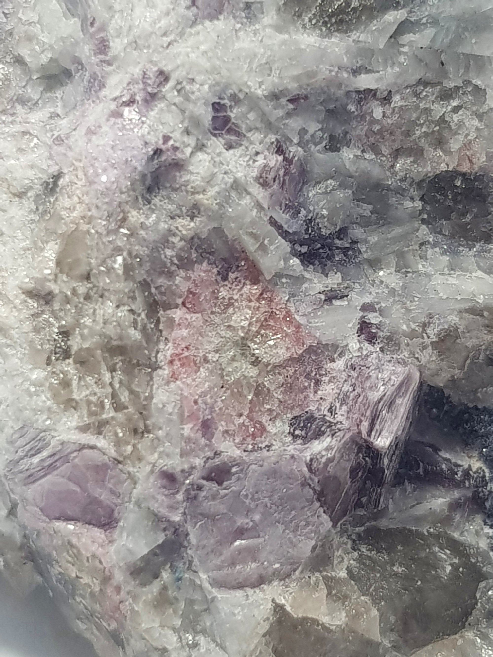 A close up view of pink tourmaline cut in cross section within a white rock.. There are multiple crystals of lepidolite. They have no clear orientation within the rock. The majority of the rock consists of awhite mineral with a blocky cleavage. Smoky quartz crystals are also present in this sample.