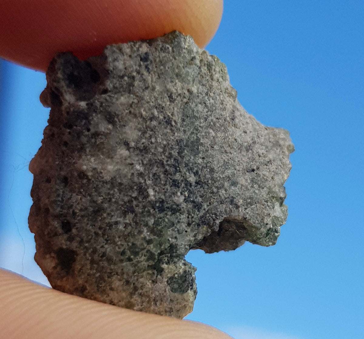 a sample of trinitite held to the sky. the piece is greenish brown, with some translucency. It does not appear to be homogenous. the surface is smoothes, the edges seem crumbly.